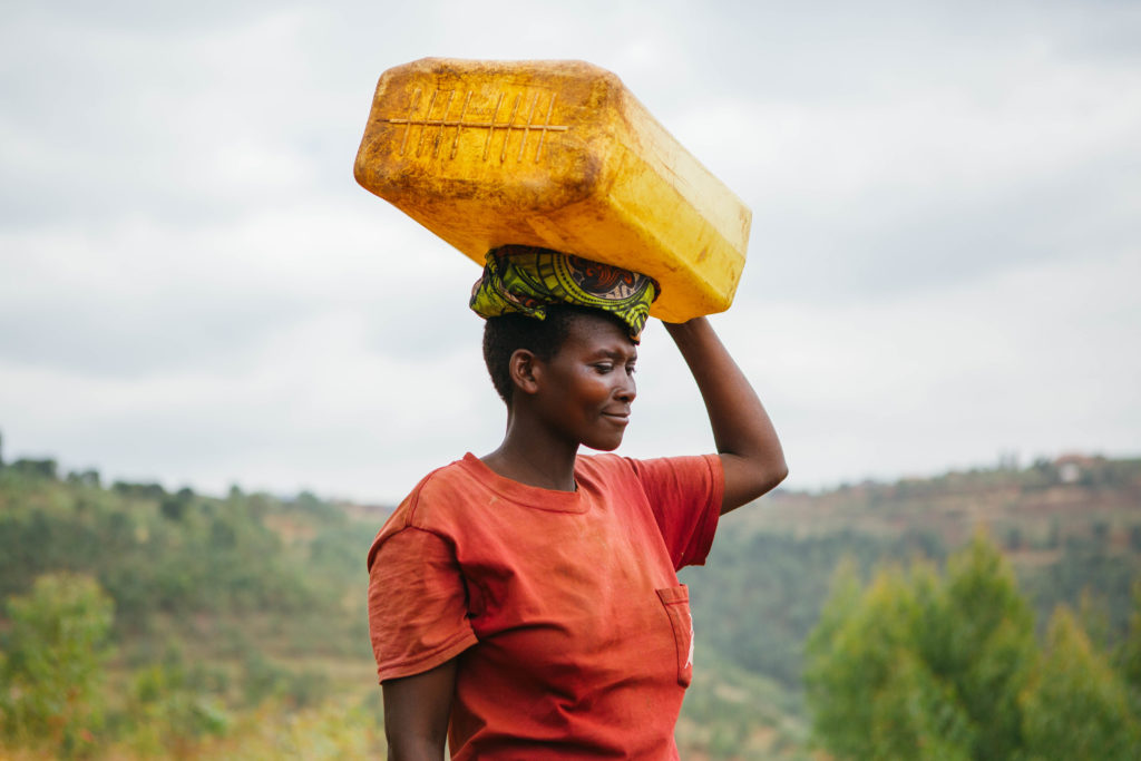 A woman in Rulindo wearing red with a mountainous background, carrying a water container. 